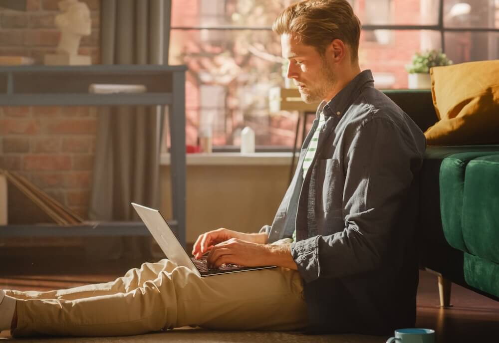 A young man is sitting on the floor and leaning against a green couch using his laptop to access Newshosting, the best Usenet provider.