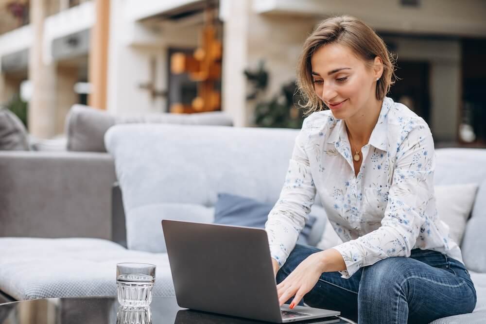 A woman is wearing a shirt with blue flowers on it and jeans. She is sitting on a couch, leaning forward, and typing on a laptop that is sitting on a coffee table.