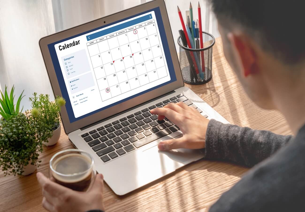 Closeup of a person’s hands typing on a laptop. They are holding an iced coffee in their left hand and are looking at a Calendar app on the screen, trying to find how far back Newshosting’s article retention goes. It’s further back than any other provider.