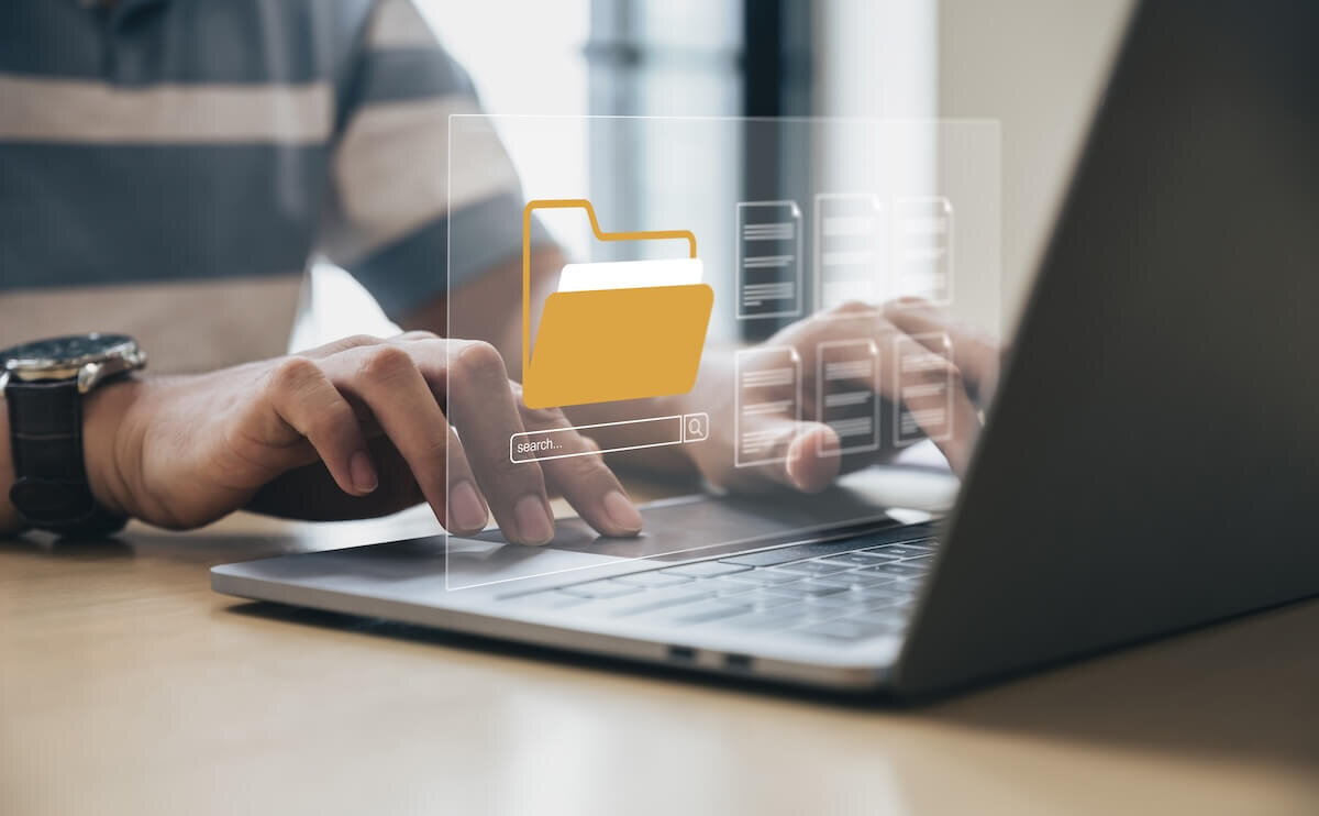 Closeup of a person’s hands typing on a laptop with a translucent window showing a yellow folder icon above a search bar and a bunch of file icons hovering above the keyboard.