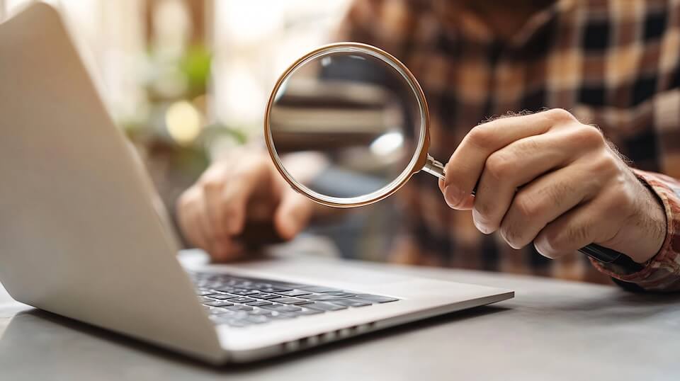 Closeup of hands near a laptop. One is hovering over a key and the other is holding a magnifying glass toward you.