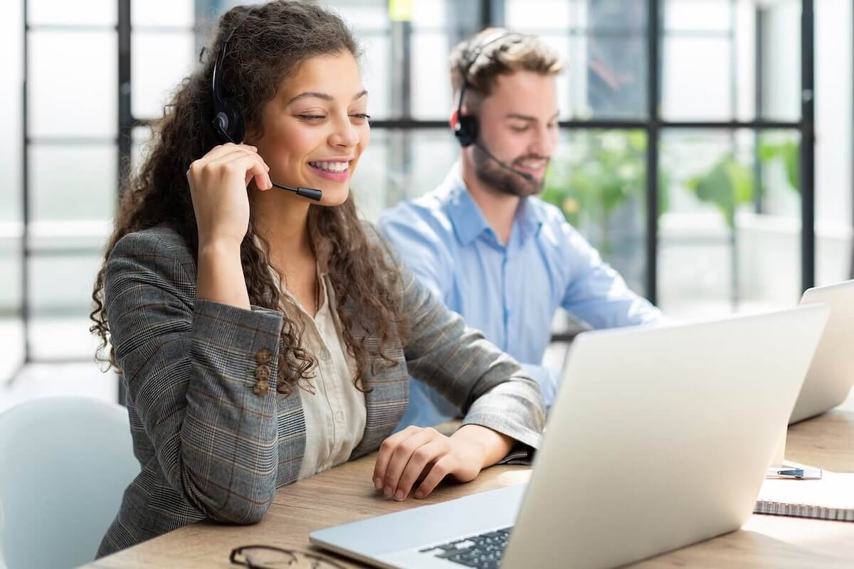A long-haired woman in a tweed coat it sitting in the foreground and a man wearing a blue button-down shirt is in the background. Both are sitting in front of laptops and wearing headsets, helping Newshosting customers.